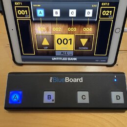 Černý MIDI kontrolér iRig BlueBoard se čtyřmi bílými tlačítky označenými písmeny A, B, C a D. Tlačítko A svítí modře. Nad ním je tablet s aplikací, k…