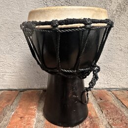 Černý perkusní nástroj typu djembe s béžovou koženou blánou. Tělo je vyrobeno z černého materiálu a horní část je stahována pleteným černým šnůrem. N…