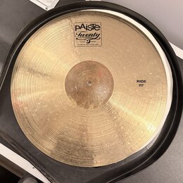 Zlatý činel s koncentrickým žilováním, umístěný v černém obalu. Na povrchu je bílý štítek s nápisy pAiste, Twenty a TRADE MARK MADE IN SWITZERLAND. V…
