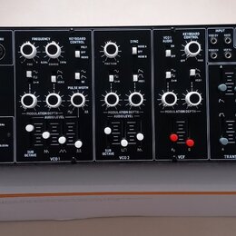 Černý elektronický syntetizátor s bílými nápisy a ovládacími prvky, umístěný na původním kartonovém obalu. Na levé straně je bílé logo behringer a ná…