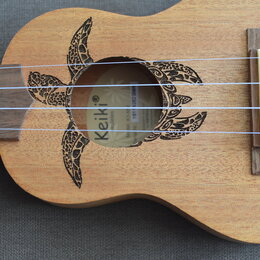 Dřevěný ukulele s vyřezaným otvorem v těle, který je zdoben vypáleným motivem želvy. Uvnitř otvoru je vidět štítek s nápisy Keiki, Ukuleles a čárový…