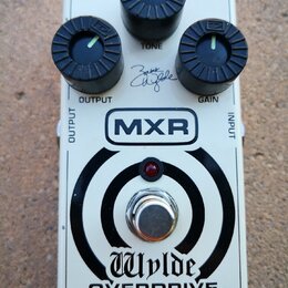 Bílý kytarový efekt pedál s nápisy MXR a Wylde OVERDRIVE. Obsahuje tři černé ovládací knoflíky s popisky OUTPUT, TONE a GAIN. Na šasi je viditelný po…