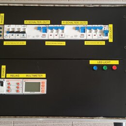 Elektrický rozvaděč v kovovém kufru s černým dnem. Horní část obsahuje jističe a štítky s nápisy PHASE, Fi SCHALTER-OUT1, Fi SCHALTER-OUT2, LED LICHT…