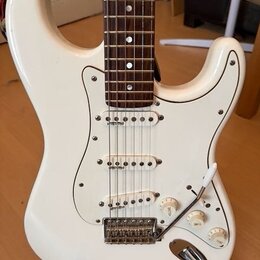 Bílá elektrická kytara typu Stratocaster s bílým pickguardem a hnědým hmatníkem. Nástroj má tři snímače typu single-coil, tři ovládací knoflíky a třy…
