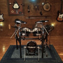 Elektronická bicí souprava značky Roland s černým rámem a chromovanými pady. Na basovém bubnu je bílý nápis Roland a pod ním menší nápis V-Drums. V p…