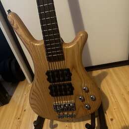 Světle hnědá elektrická basová kytara s přírodným dřevem a viditelnou masivní strukturou zrna. Nástroj má dva černé humbuckery, čtyři ovládací knoflí…