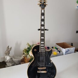 Černá elektrická kytara typu Les Paul s bílými hrankami a zlatým hardwarem. Na hlavě je nápis Epiphone a logo v podobě křídla. Kytara má dva humbucke…