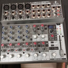 Stříbrný mixážní pult s nápisem BEHRINGER XENYX 1202FX. Zařízení obsahuje několik vstupních kanálů s potenciometry, přepínači a konektory typu XLR a…