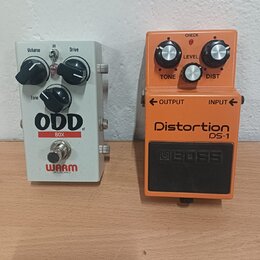 Dvě kytarové efektní pedály umístěné vedle sebe na dřevěném povrchu. Vlevo je bílý pedál s nápisy ODD BOX v1 a WARM AUDIO, vybavený třemi černými kno…