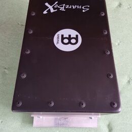 Černý obdélníkový box s nápisem SnareBox a bílým kulatým logem MEINL. Horní kryt je přišroubován několika černými šrouby. V dolní části je vidět kovo…