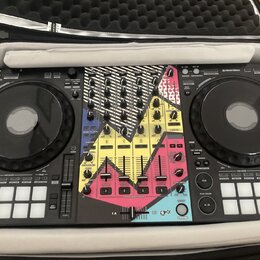 Černý DJ 컨트롤러 umístěný v přepravním kufru. Střední část přístroje je pokryta barevnou polepkou s růžovými, žlutými a modrými geometrickými tvary. V p…