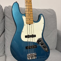 Modrá elektrická basová kytara s bílým pickguardem a hnedým javorovým hmatníkem. Nástroj má dva snímače a ovládací panel s třemi knoflíky. Na těle ne…