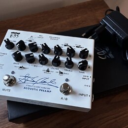 Bílý akustický předzesilovač s nápisem EBS a Stanley Clarke Signature ACOUSTIC PREAMP. Zařízení má několik ovládacích knoflíků, přepínačů a vstupů/vý…