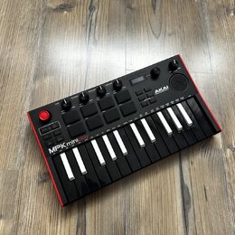 Černý kompaktní MIDI kontrolér s červeným rámem, umístěný na dřevěné podlaze. Na horní části je nápis AKAI PROFESSIONAL a v dolní části model MPKmini…