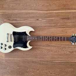 Bílá elektrická kytara s černým plechtrátem a hnědým hmatníkem. Na hlavě kytary je bílý nápis Epiphone a pod ním označení SG. Nástroj vykazuje mírné…