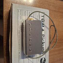 Krytovaný magnetický snímač stříbrné barvy s pěti viditelnými šrouby. Snímač je připojen k opletenému kabelu a položen na kartonu s nápisy TUMA Picku…