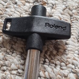 Černý plastový klíč v tvaru kříže s kovovým hrotem. Na plastové části je vyražen nápis Roland. Předmět leží na světlém koberci.