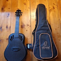 Černá kytara s povrchem imitujícím karbonové vlákno a doprovodný černý pouzdro s oranžovým lemováním. Na pouzdře je bílé logo a nápis ORTEGA GUITARS…