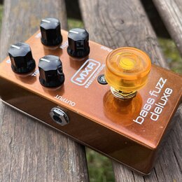Bronzový kytarový efekt s nápisy MXR, BASS INNOVATIONS a bass fuzz deluxe. Zařízení má čtyři černé plastové knofíky a jeden velký průsvitný oranžový…