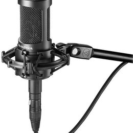 Černý kondenzátorový mikrofon umístěný v odpruženém držáku na černém stativu. Na těle mikrofonu je bílý nápis audio-technica. Na spojovací části stat…