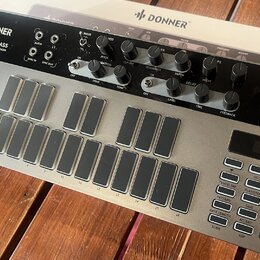 Stříbrný analogový syntetizátor s černým ovládacím panelem. Na horní části je nápis DONNER SYN BASS ANALOG SYNTHESIZER AND SEQUENCER a logo DONNER. P…