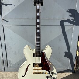 Kytara typu semi-akustika v krémové barvě s hnědočerveným pickguardem a zlatými komponenty. Na hlavě je bílý nápis Epiphone. Hриф má vložky z perlece…