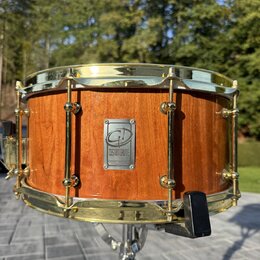 Dřevěný buben s přirozeným povrchem a zlatými kovovými obručemi. Uprostřed korpusu je kovový štítek s logem a nápisem CUSTOM DRUMS. Nástroj vykazuje…