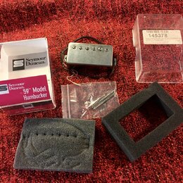 Na červeném povrchu je položen kytarový snímač typu humbucker v kovovém krytu s černými vodiči. Vedle něj je bílá krabička a červený papírový obal s…