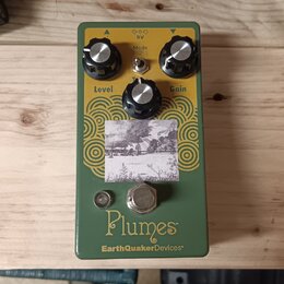 Zelený kytarový efekt s nápisy Plumes a EarthQuakerDevices®. Na horní straně jsou tři černé kontrolní knoflíky s popisky Level, Tone a Gain a přepína…