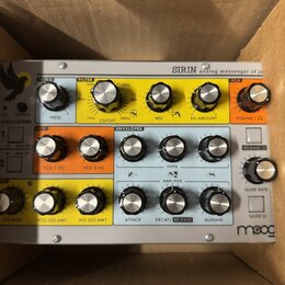 Stříbrný analogový syntetizátor Moog Sirin umístěný v kartónovém boxu. Přední panel obsahuje množství ovládacích knoflíků rozdělených do barevných se…