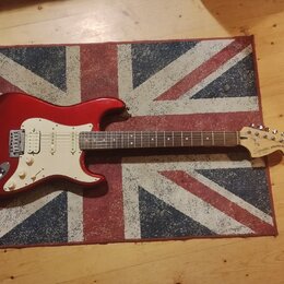 Červená elektrická kytara s bílým pickguardem a hnědým hmatníkem. Na hlavě je viditelné logo Squier STRATOCASTER. Nástroj leží na koberci s motivem b…