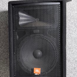 Černý reproduktor v bedně potažené látkou s kovovou mřížkou. V dolní části je oranžový čtverečkový štítek s bílým nápisem JBL a pod ním menší text JR…