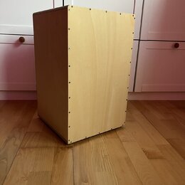 Světle hnědý, dřevěný reproduktorový box s hranatým tvarem. Boční stěna je spojena šrouby viditelnými v řadě podél hran. Povrch dřeva je hladký bez v…