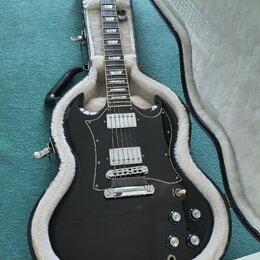 Černá elektrická kytara s dvojstupňovými humbuckery a bílým pickguardem, umístěná v otevřeném černém kufru s bílou vnitřní výstylou. Na hlavě nástroj…