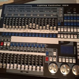 Černý světelný kontrolér s nápisem Lighting Controller 1024. Zařízení obsahuje množství posuvníků, tlačítek a tři velké stříbrné otočné ovladače v do…