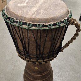 Djembe s tělem z tmavého dřeva a koženou blánou. Horní okraj je opleten zeleným šňůrkem a nástroj je napnutý černými provazníky. Na pravé straně je p…