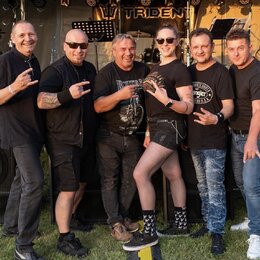Fotografie zobrazuje skupinu šesti lidí v černém oblečení před pódiem s nápisem TRIDENT. V pozadí jsou vidět reproduktory, osvětlovací technika a čás…