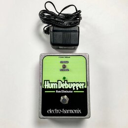 Kytarový pedál značky electro-harmonix s názvem Hum Debugger a přívostkem Hum Eliminator. Zařízení má stříbrný kovový šasi, zelený vršek s vodorovným…