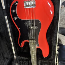 Červená elektrická basová kytara v černém pouzdře. Nástroj má černý pickguard a dvě kontrolní knoflíky. Na kytaru nejsou viditelné žádné značky ani n…