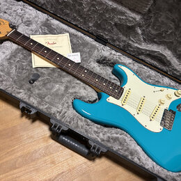 Modrá elektrická kytara typu Stratocaster s bílým pickguardem a hnědým hmatníkem, položená v šedém tvrdém kufru. Na hlavě kytary je viditelné logo Fe…