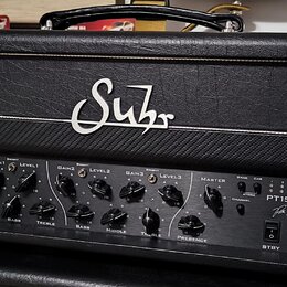 Kytarový zesilovač v černém koženěném potahu s bílým lemováním. Na horní části je bílé logo "Suhr". Přední panel obsahuje ovládací knoflíky pro Gain,…