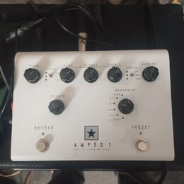 Bílý kytarový pedál s nápisem AMPED 1 a pod ním menším textem DEPT. 10 | 100W AMP PEDAL. Nad nápisem je logo ve tvaru hvězdy v černém čtverci. Zaříze…