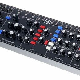 Černý modulární syntetizátor v dřevěném šasi s bílým spodním panelem. Na horním panelu jsou rozděleny sekce s názvy CONTROLLERS, OSCILLATOR BANK, MIX…
