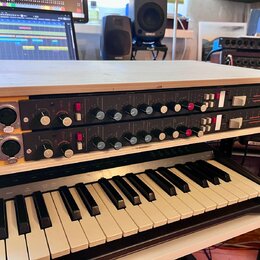 Dva identické rackové audio procesory v černé barvě, umístěné v bílém dřevěném šasi s žlutým bočním panelem. Každý jednotka má několik otočných knofl…