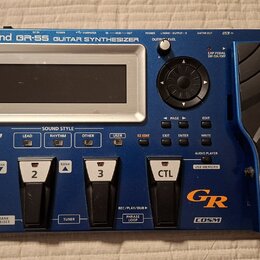 Modrý kytarový syntetizátor s nápisem Roland GR-55 GUITAR SYNTHESIZER. Zařízení má displej, ovládací knoflík se šipkami a čtyřistupňové pedály označe…