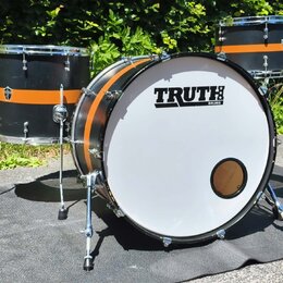 Černá bubnová sada s oranžovými pruhy, která obsahuje basový buben a dva tom-tomy. Na bílém předním zrcadle basového bubnu je černý nápis TRUTH DRUMS…