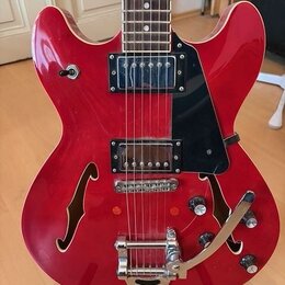 Červená polodupatá elektrická kytara s černým plechtrátem. Nástroj má dva humbuckery, dva f-otvory a vibrační systém s páčkou. Na těle nejsou viditel…