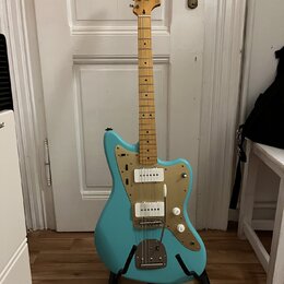 Modrá elektrická kytara s jasným dřevěným hřidlem a zlatým pickguardem. Na hlavě je viditelné logo "Supro". Kytara má dva humbuckery, dvě ovládací kn…