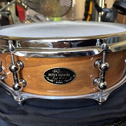 Dřevěný buben s přirozeným povrchem a chromovanými obručemi. Na korpusu je oválný černý štítek s nápisy „PETER ERSKINE“, „Jazz Snare“ a logem „TAMA“.…