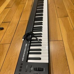 Černý klaviaturní kontrolér s bílými a černými klávesami, položený na dřevěné podlaze. Na levé straně je nápis KEYSTATION 88 MK3 a v horní části logo…